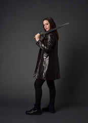 full length portrait of brunette girl wearing long leather coat and boots. standing pose and holding a sword on grey studio background.