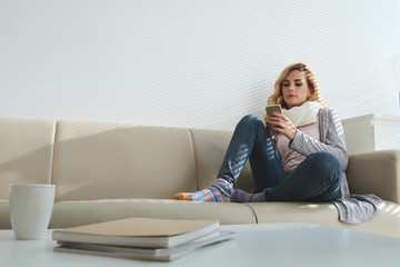 Attractive young in scarf and warm clothes resting on sofa and texting friends