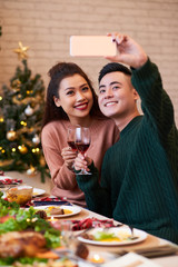 Cheerful young Asian couple photographing together at Christmas dinner table