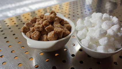 Brown and white sugar cubes in ceramic bowls on a metal counter with holes in a restaurant