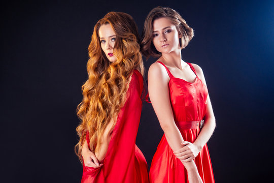 fashion photo of two beautiful women with luxurious long dark hair and short haircut in a red silk dress posing in studio on black background.perfect skin, professional makeup sensual model sisters