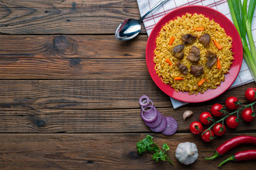 Red plate with pilaf on a brown wooden table. On the table is red pepper, green onions, garlic, cherry tomatoes, red napkin, spoon. Top view. Flat lay.