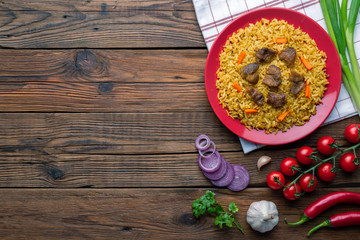 Red plate with pilaf on a brown wooden table. On the table is red pepper, green onions, garlic, cherry tomatoes, red napkin, spoon. Top view. Flat lay.