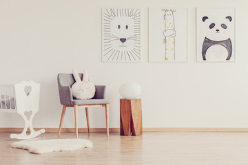 Grey chair with pink pillow between wooden stool and white crib in scandinavian baby room interior with posters on the wall © Photographee.eu