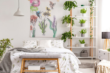 Real photo of white bedroom interior with many fresh plants, king-size bed, material painting with floral pattern and bench with books