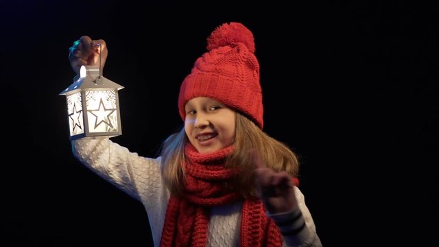 Little girl wearing knitted winter hat and scarf standing with lantern searching looking to sides and gesturing no with a finger at the end, over black background