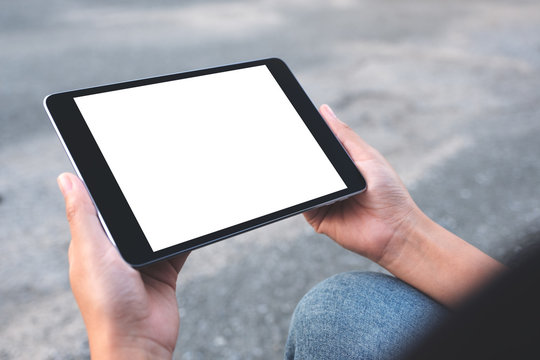 Mockup image of a woman holding black tablet pc with blank white screen while sitting on the street background