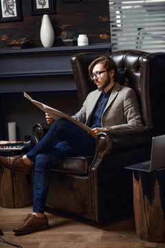 A Man In A Wooden Office Sits In A Leather Armchair With A Newspaper. Left View.