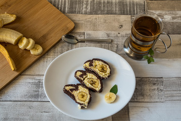 Banana cake with chocolate sauce and cup of tea on a white plate with wooden background