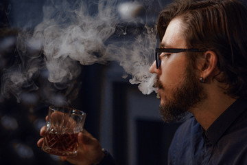 Long-haired hipster emits thick cigarette smoke. Man holding a glass with alcohol