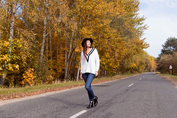 Fototapeta premium Red-haired girl in a white sweater and a black hat is on the road in the autumn forest