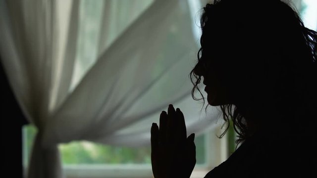 Woman silhouette praying indoors, reading a pray with hands folded, asking God