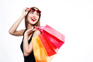 Sexy brunette woman wearing black dress and sunglasses standing with colorful shopping bags and looking up on the white background, concept of consumerism, sale, rich life