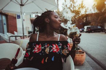 Portrait of a charming African girl sitting in a street cafe and making smoke in the glass of...