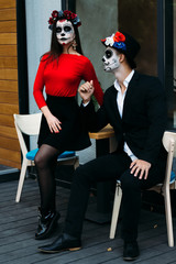 A couple, wearing skull make-up for. All souls day. Boy and girl sugar skull makeup.painted for halloween sit in a cafe. dead in the city. zombie walk.