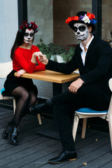 Fototapeta na wymiar A couple, wearing skull make-up for. All souls day. Boy and girl sugar skull makeup.painted for halloween sit in a cafe. dead in the city. zombie walk.