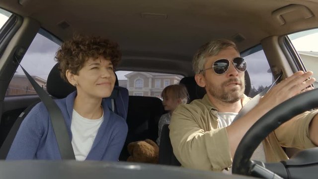 Shot from inside of moving car: bearded man in sunglasses driving as cheerful woman with short hair chatting and pointing at something out window, then looking at little girl and boy sitting in back