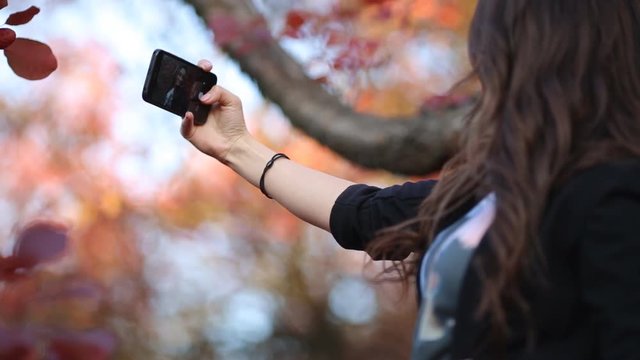 A charming young girl takes pictures of herself on a mobile phone. Brunette with a beautiful smile in the autumn garden takes a selfie on a smartphone.