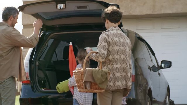 PAN shot of cheerful family with children packing for picnic: they are loading rucksack, blanket, umbrella and basket into trunk of car, then entering vehicle