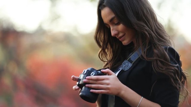 A charming young girl photographer shoots the autumn landscape with a digital SLR camera. A brunette with a beautiful smile in the autumn garden takes pictures of trees with red leaves.