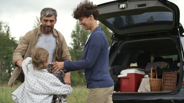 PAN shot of happy mother with short hair, father with beard and cute little girl folding blanket and putting it in trunk of car after picnic in park