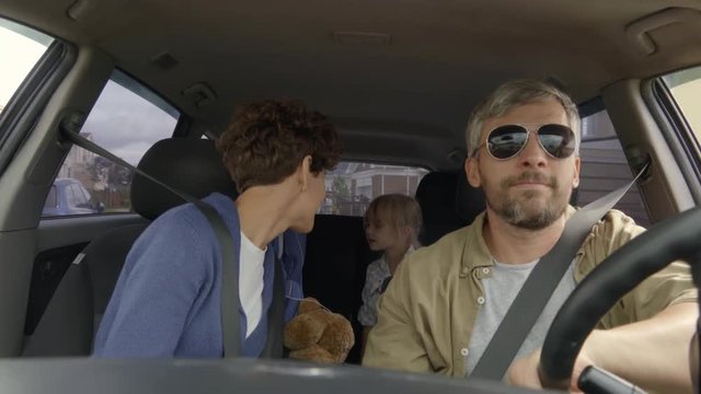 Shot from inside of moving car: bearded man in sunglasses driving as happy woman with short hair sitting in passenger seat chatting with little girl and boy sitting in back