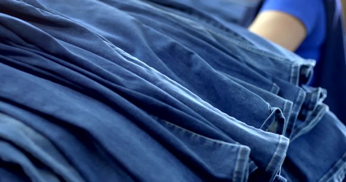 Focus on piles of folded jeans with unidentifiable female garment industry worker sorting clothes in background at warehouse in Los Angeles. Slow motion hand-held 4K recorded 60fps