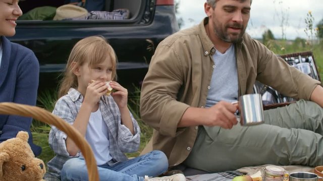 PAN shot of cute little girl sitting cross-legged on blanket and enjoying sandwich while having picnic with happy mother with short hair and bearded father drinking hot tea from mug