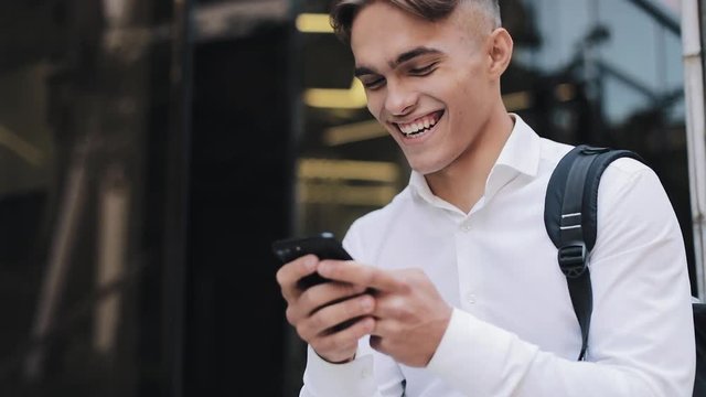 Happy young businessman or tourist with a backpack using phone sitting near modern office building. Communication Successful Rich Portrait Modern Handsome Smartphone Ambitious