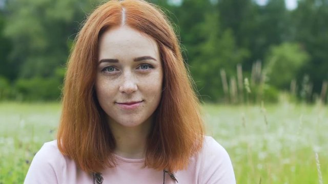 Red haired woman with freckles looks in camera outdoors in field, female smile