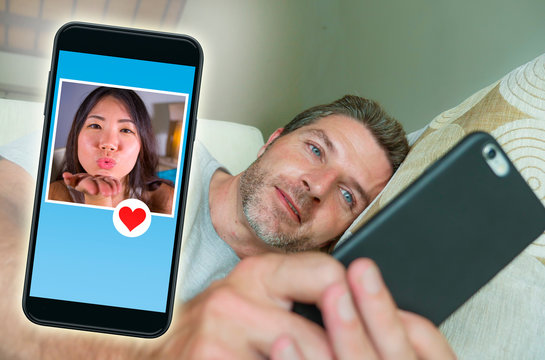 young handsome and happy man using internet social media on mobile phone searching love flirting and dating online sending like to beautiful Asian Chinese woman