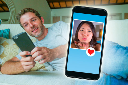 young handsome and happy man using internet social media on mobile phone searching love flirting and dating online sending like to beautiful Asian Chinese woman