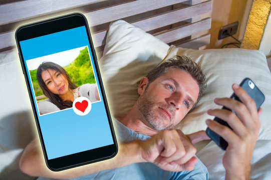 young handsome and happy man using internet social media on mobile phone searching love flirting and dating online sending like to beautiful Asian Chinese woman