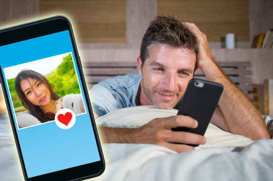 young handsome and happy man using internet social media on mobile phone searching love flirting and dating online sending like to beautiful Asian Chinese woman