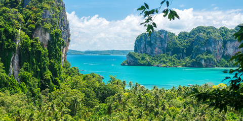Railay, Thailand: cliffs and rocks overgrown with jungles, coconut palm trees and a beautiful bay with plying boats in the distance during a sunny day. Evergreen tropical paradise in Krabi province