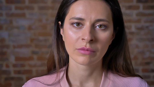 Sad face of caucasian woman looking at camera close-up and not blinking while standing near red brick wall indoor, casual pink jacket