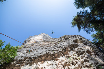 From below shot of climber secured with rope jumping from top of mountain under blue sky while other is climbing 