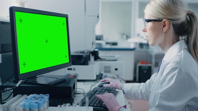 Female Research Scientist Sits at His Workplace in Laboratory, Uses Green Mock-up Screen Personal Computer. I the Background Genetics, Pharmaceutical Research Centre.