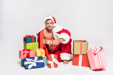 sexy man in santa costume sitting with gift boxes and smiling at camera on grey