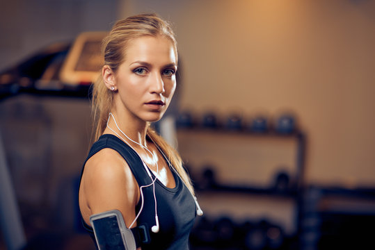 Close up of woman standing in gym and looking in camera. Around neck earphones. Healthy lifestyle concept.