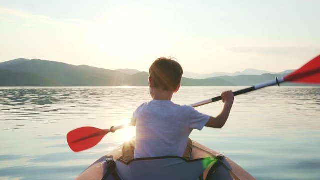 Slow motion of a happy boy with his dog Jack Russell Terrier paddling an inflatable kayak on the water of a large mountain lake against the backdrop of beautiful orange sunset. Family sports vacation