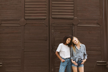 Young nice african american woman with dark curly hair in T-shirt and jeans and cute woman with blond hair in shirt and shorts joyfully looking in camera with brown wall on background