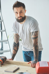 handsome bearded man leaning at wooden table and looking at camera during house repair
