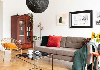 Table in front of armchair and sofa in white flat interior with wooden cabinet and posters. Real...