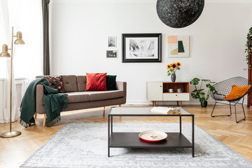 Table on carpet in white apartment interior with sofa, armchair and posters above cabinet. Real...