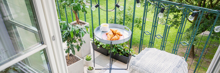 Croissants and tea on the table on bright balcony with pouf and green plants in pots, panoramic...