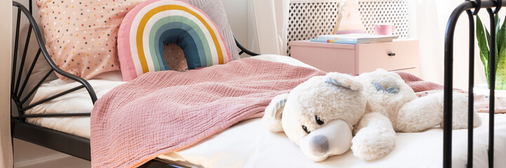 Close-up of child bed with teddy bear, pastel pink blanket and rainbow cushion in the real photo