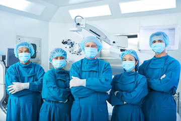 Asian surgical team in masks and uniform keeping arms crossed and looking at camera while standing...