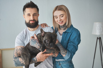 happy young couple with tattoos holding cute french bulldog in new home