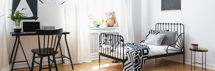 White teenager room interior with books and laptop on desk, window with drapes and metal bed with black and white sheets